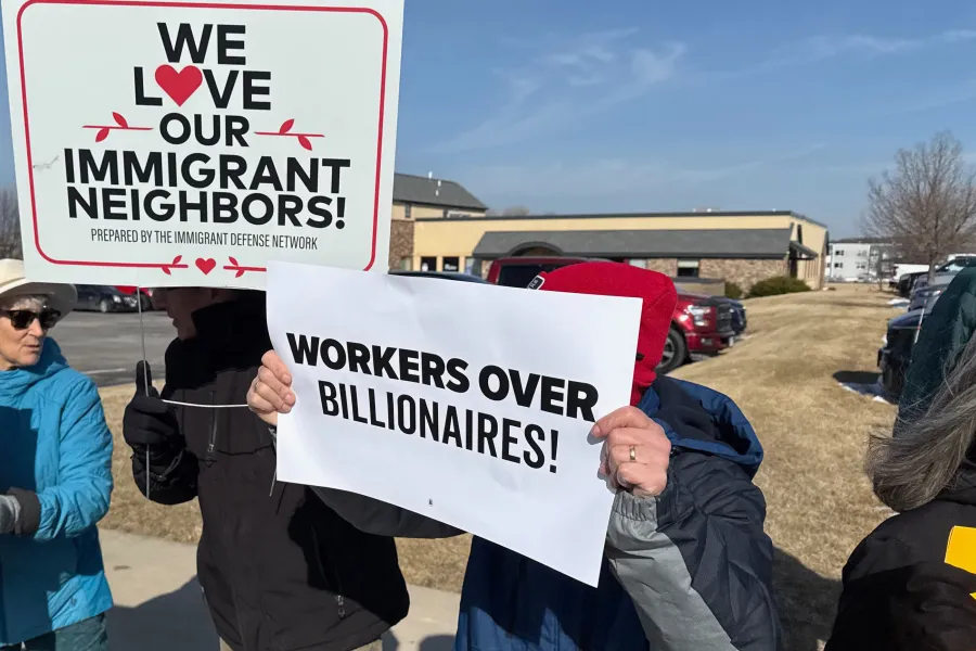 People holding signs. One reads "We Love our Immigrant Neighbors." The other reads "Workers over billionaires!"